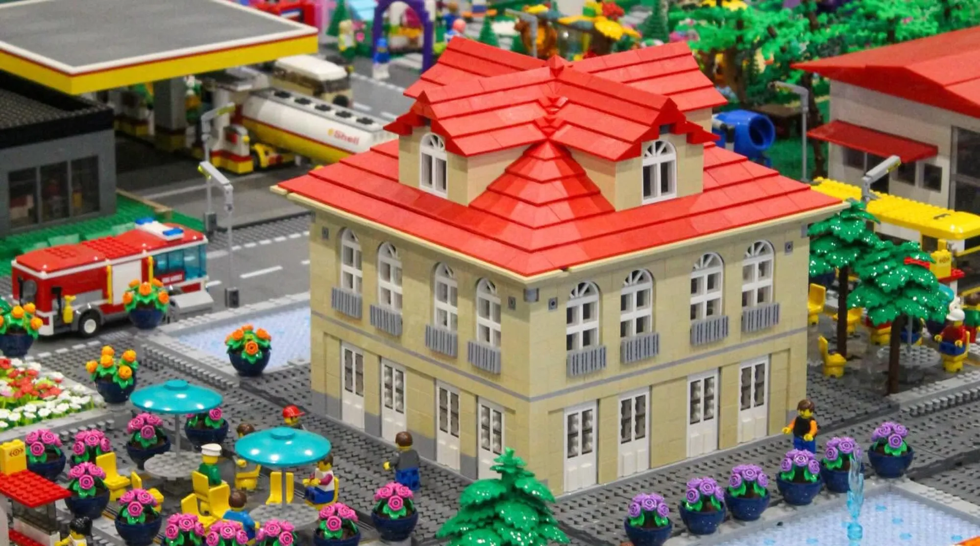LEGO diorama featuring a cream-colored building with red roof, minifigures, flower planters, and surrounding structures in…