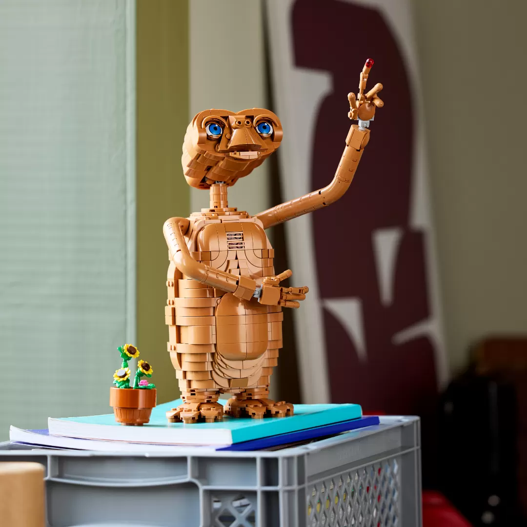 LEGO E.T. figure with glowing blue eyes and outstretched finger, displayed on books with a small potted flower beside it.