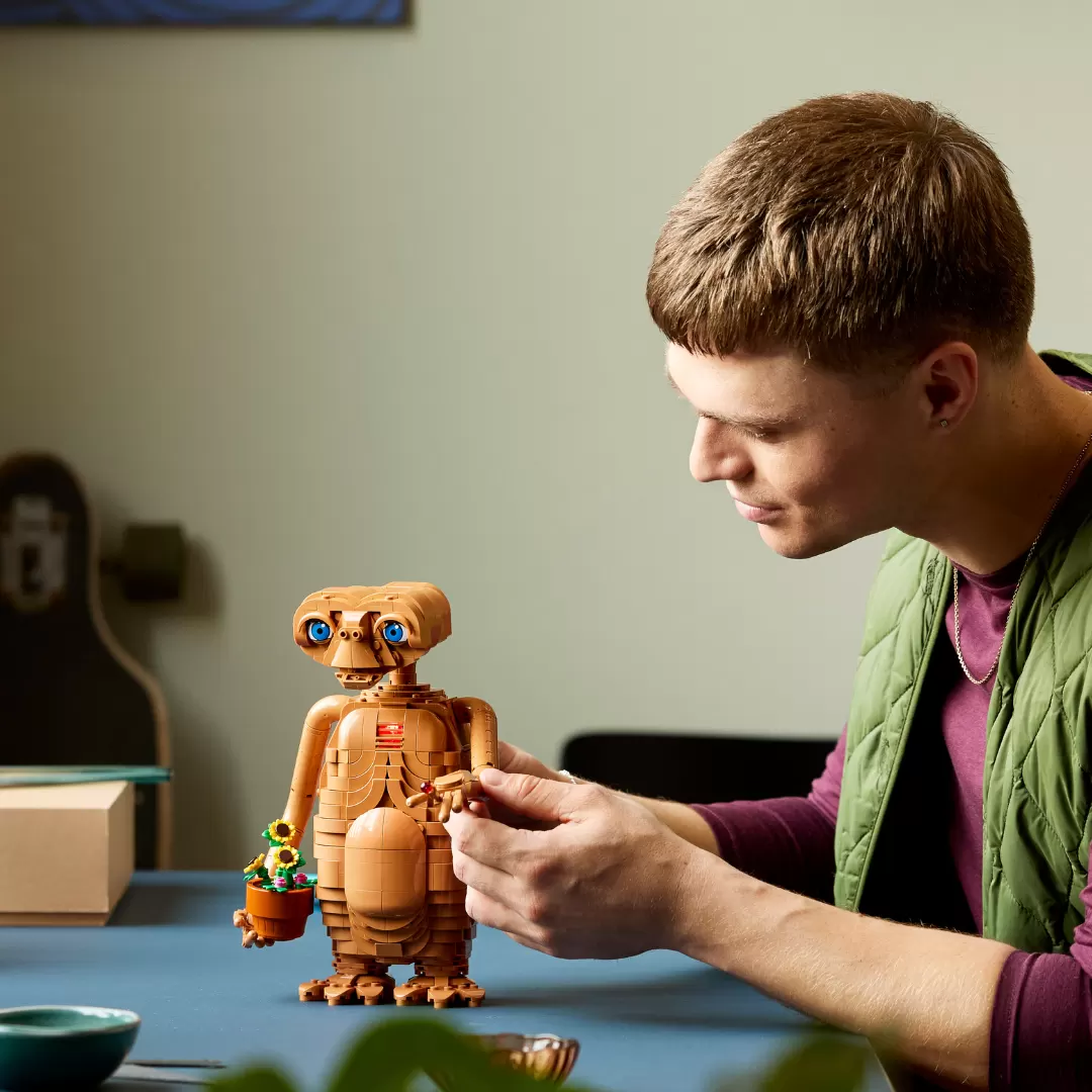 Young man examining a tan LEGO E.T. figure with glowing blue eyes and a small potted plant accessory at a table.