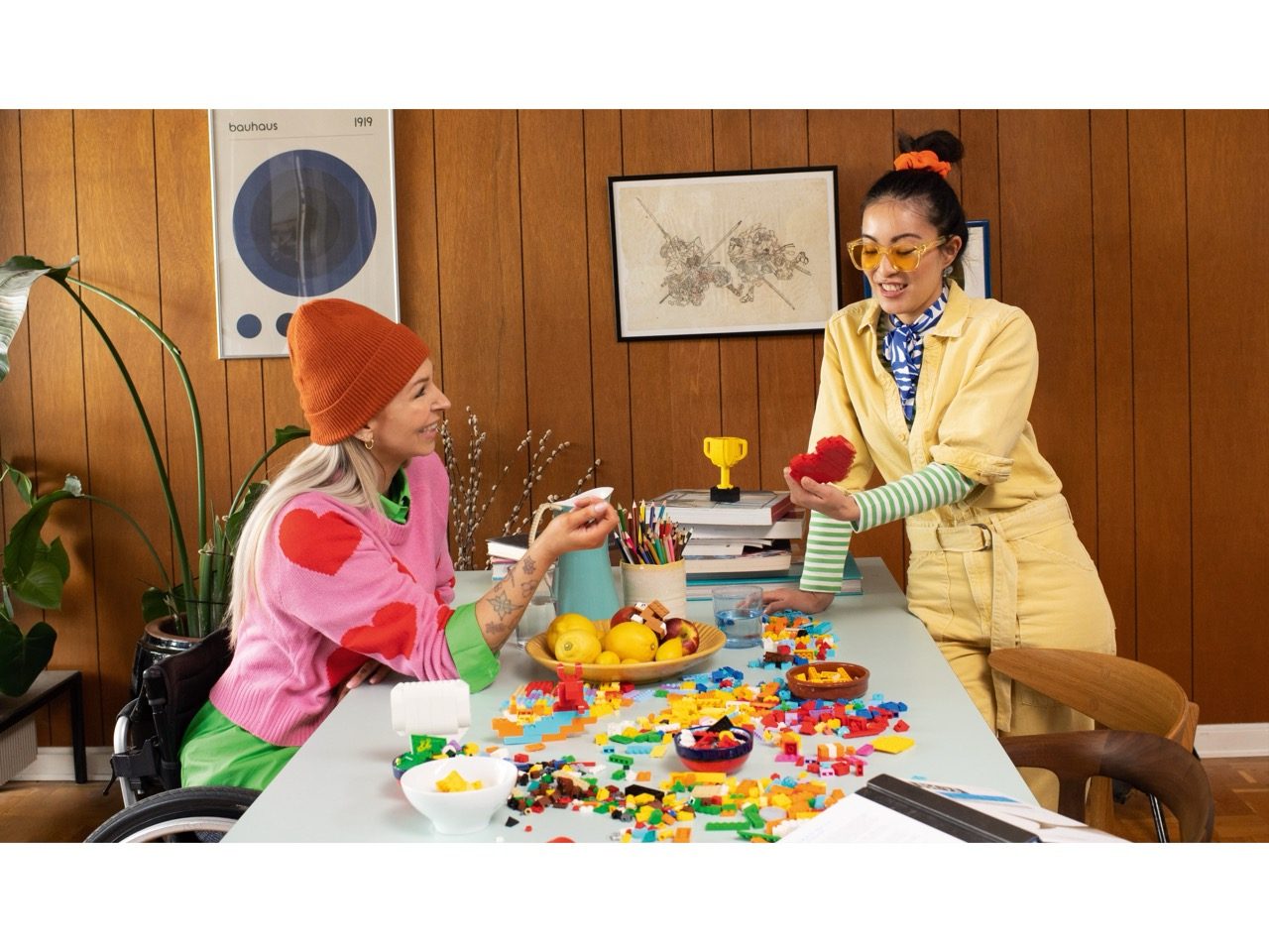 Two women at a desk with colorful LEGO bricks, minifigures, and accessories in a retro wooden office setting.