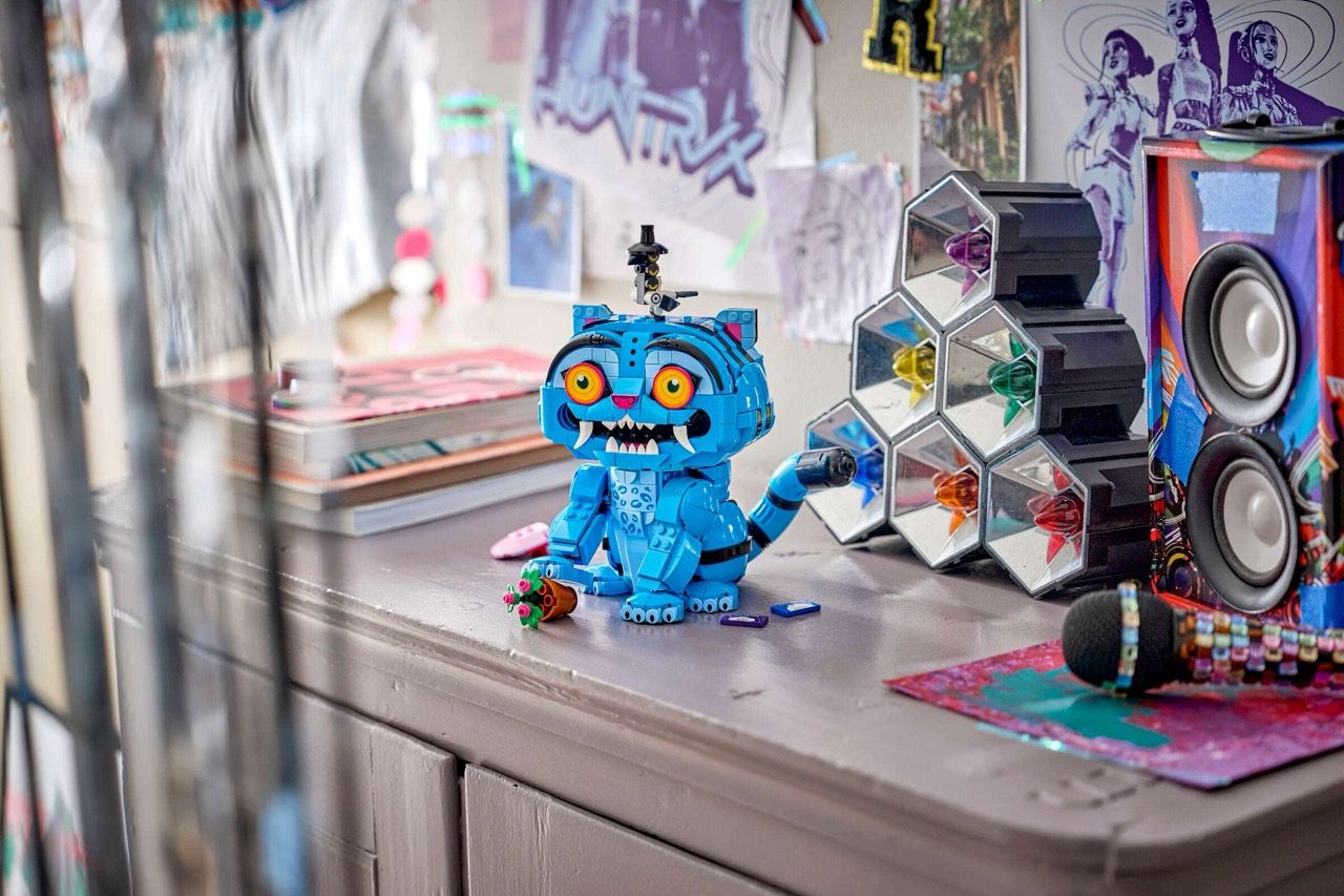 Blue LEGO cat figure with yellow eyes and grinning teeth displayed among colorful accessories on a shelf.
