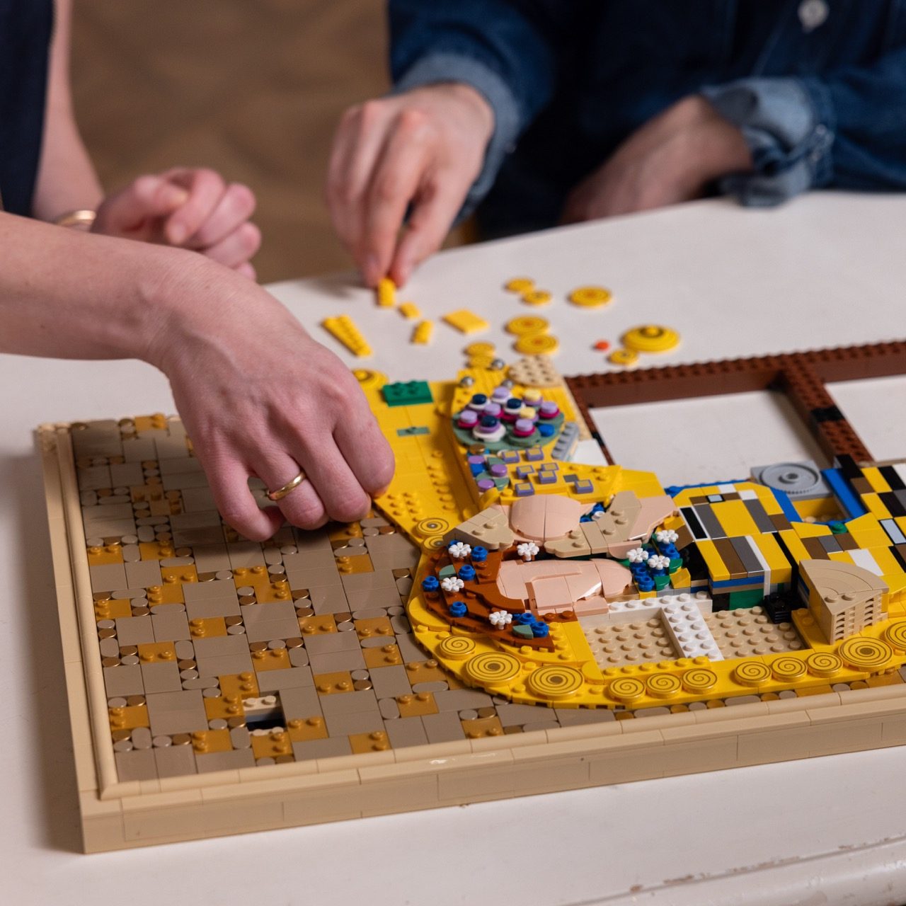 LEGO Art Gustav Klimt set with gold and colorful tiles being assembled by multiple hands on a white table.