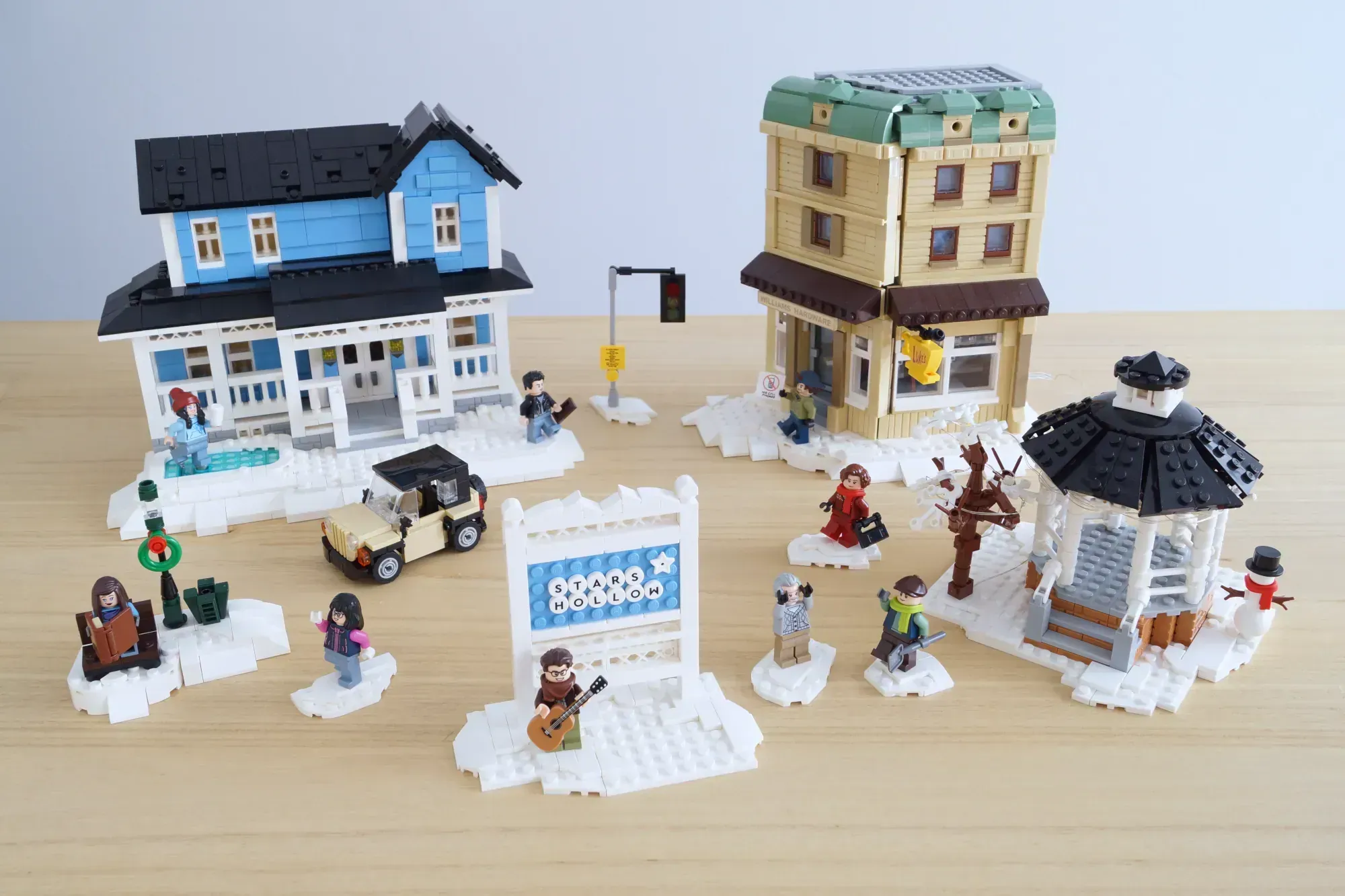 LEGO winter village scene with blue house, tan building, gazebo, minifigures, vehicle, and snow bases on wooden surface.