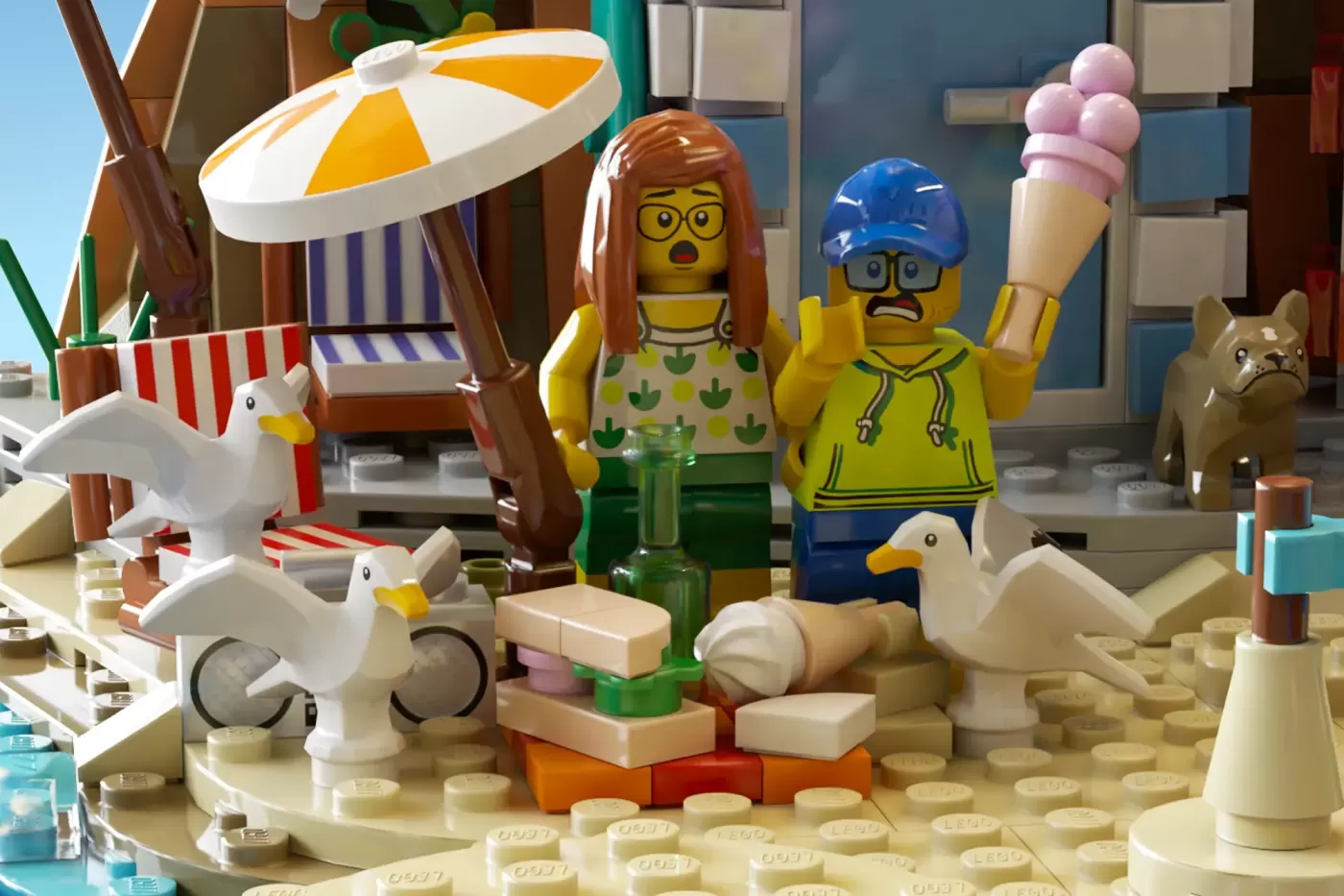 LEGO seaside pier scene with two minifigures, seagulls, beach umbrella, ice cream cone, and Victorian-style buildings.