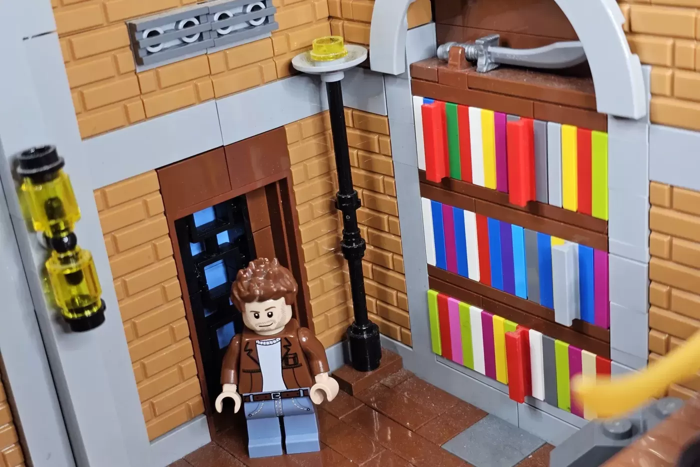 LEGO minifigure in brown jacket standing in brick library with colorful books on shelves and a black lamp post.