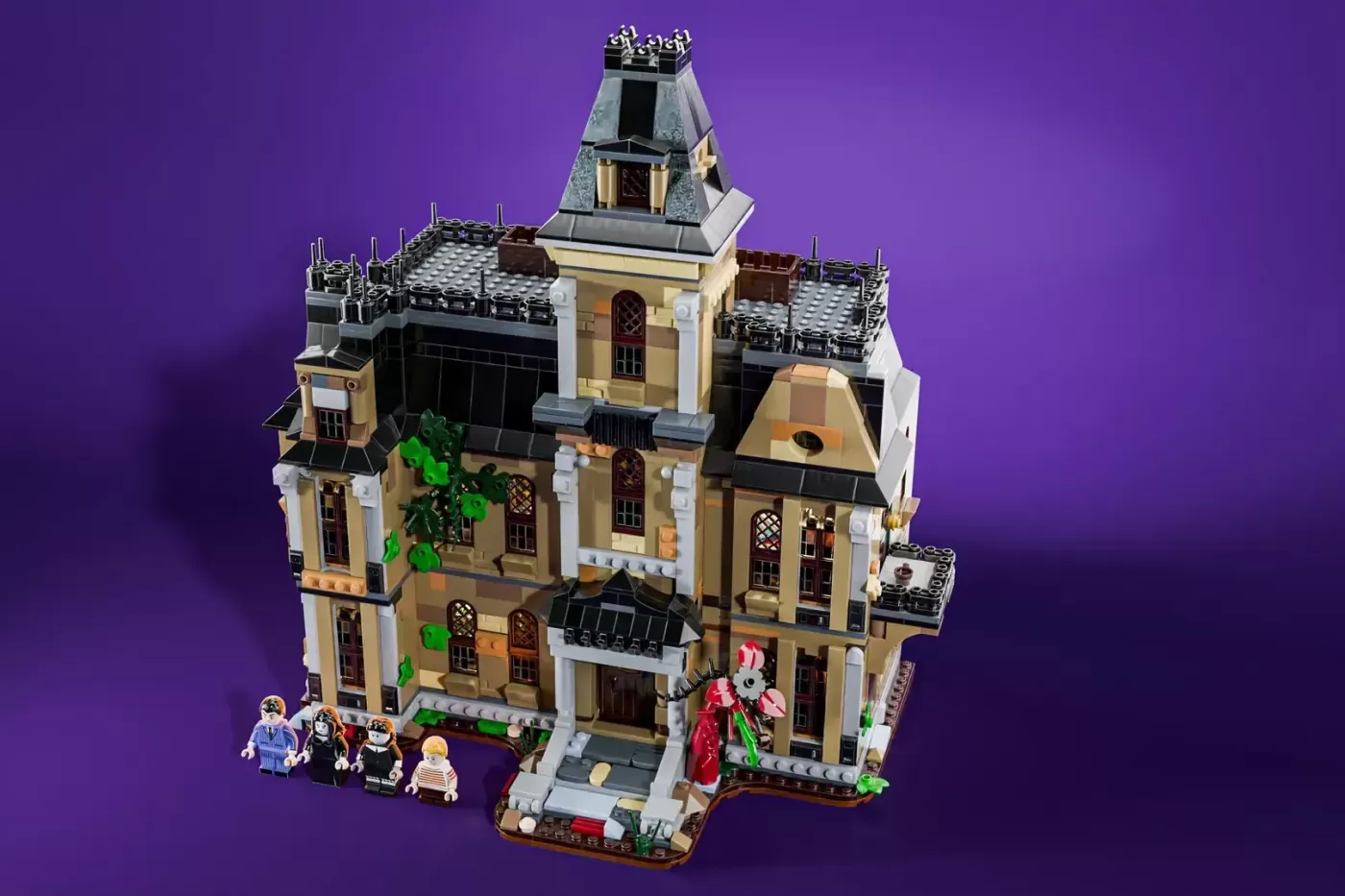 LEGO Addams Family Mansion set with detailed architecture, tower, minifigures, and decorative plants against purple backgr…